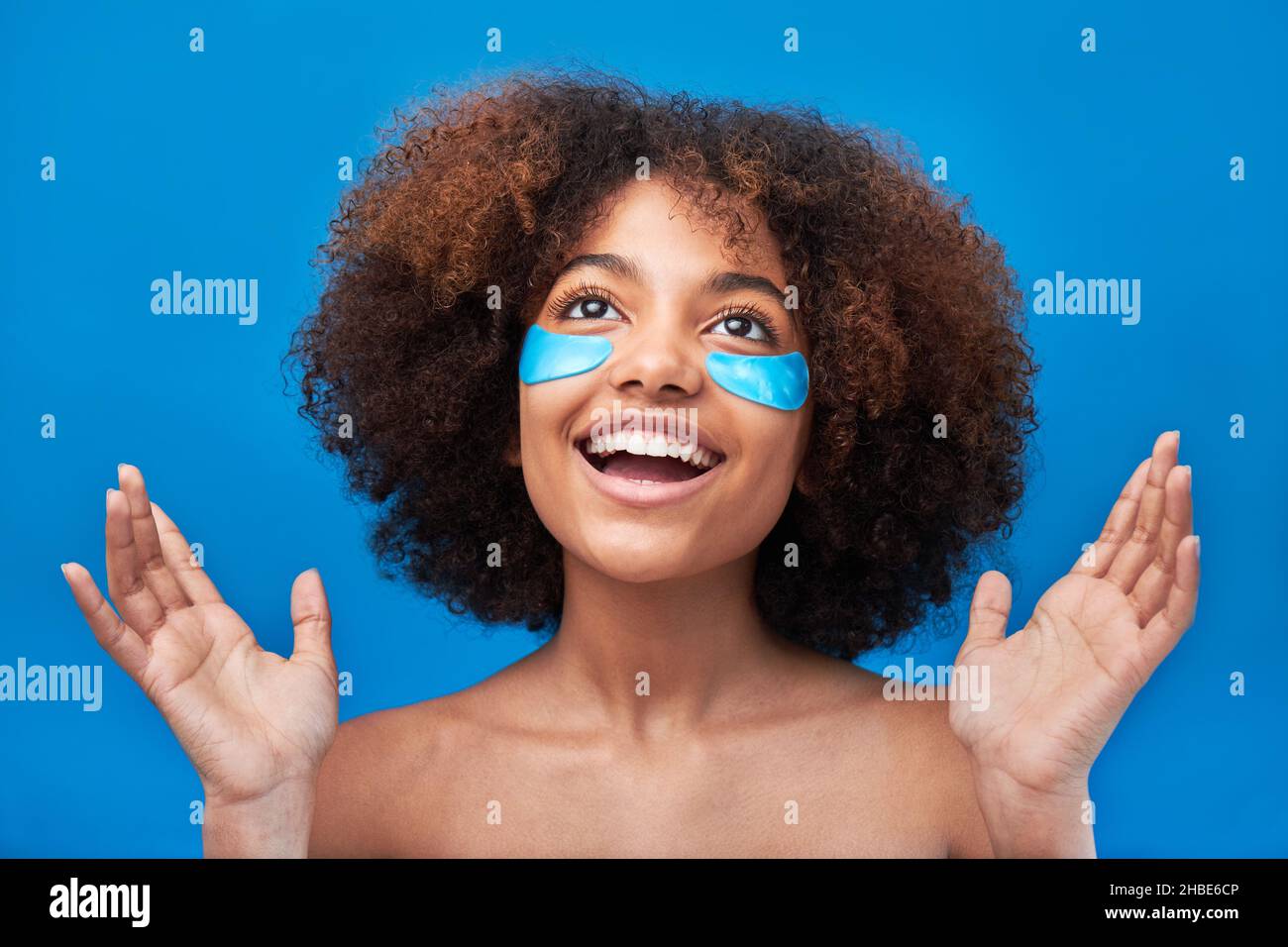 Junge Frau mit langen lockigen Brünetten Haaren posiert mit breitem Lächeln und blauen Flecken unter den Augen gegen helle Studiowand Nahaufnahme Stockfoto
