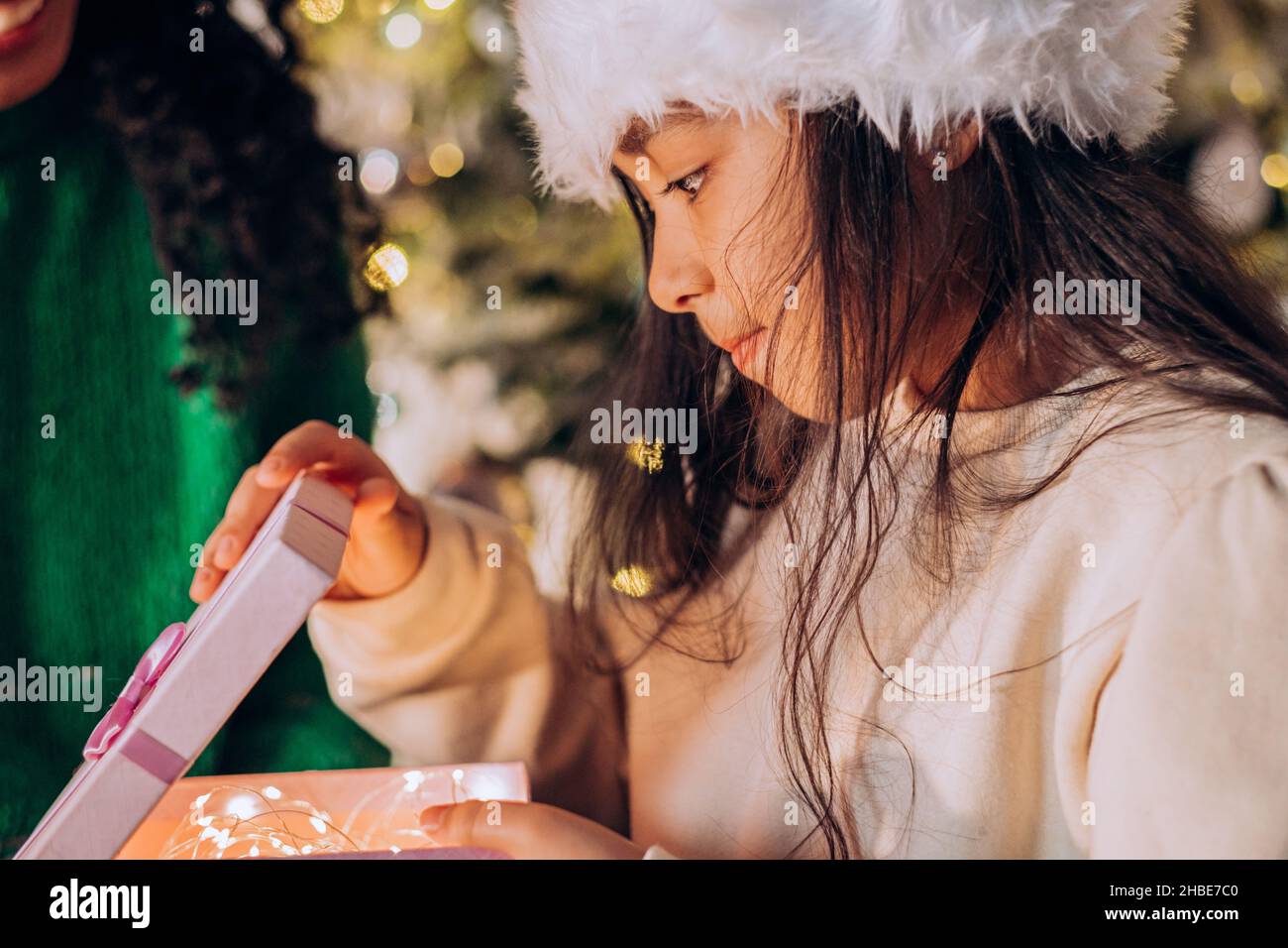 Brünette langhaarige Tochter in Santa Hut öffnet festliche Box mit Geschenken sitzen in der Nähe afroamerikanische Mutter gegen Weihnachtsbaum Stockfoto