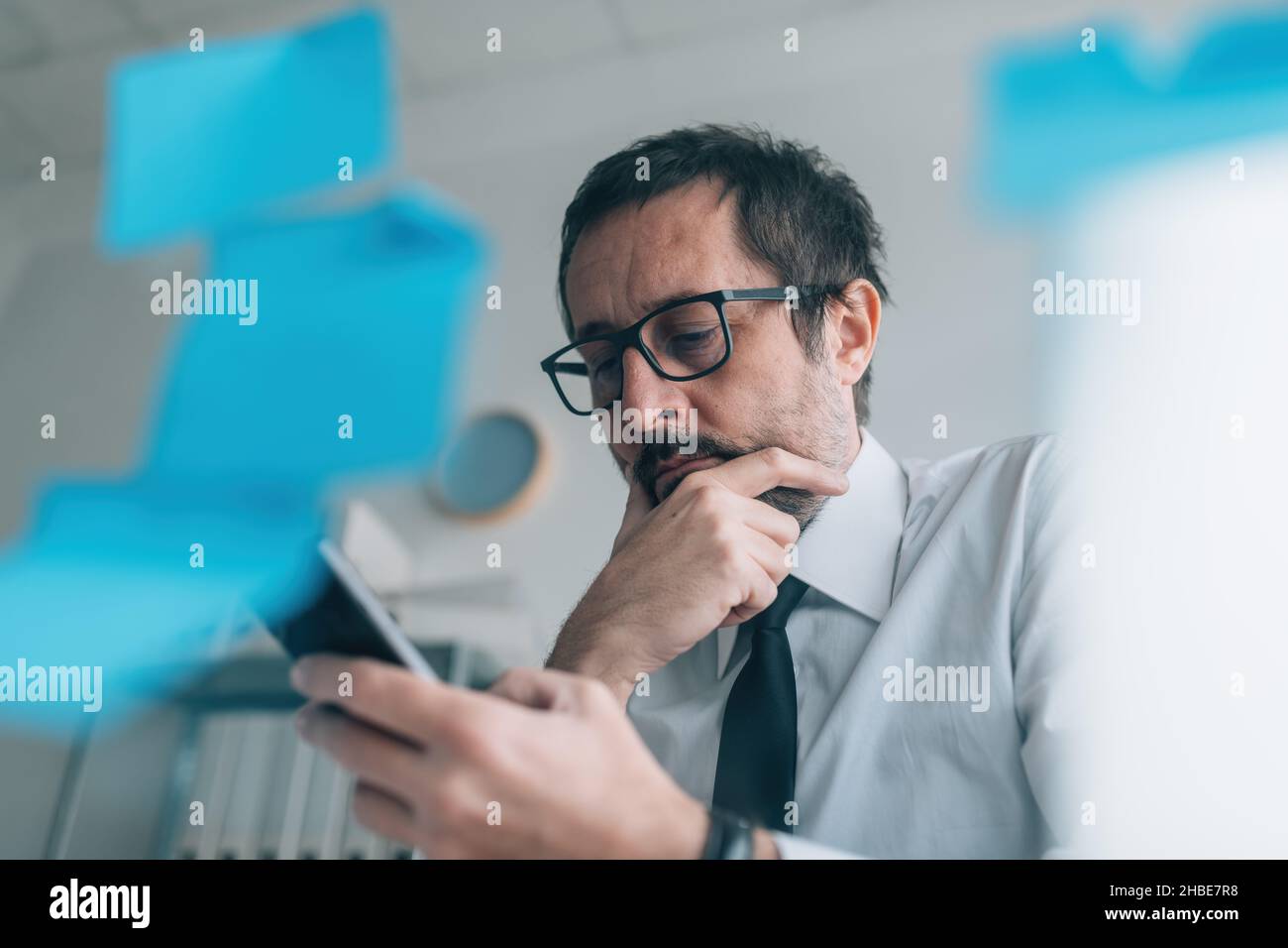 Rücksichtsvoller Geschäftsmann, der an eine SMS-Antwort denkt und das Mobiltelefon im Büro ansieht, selektiver Fokus Stockfoto
