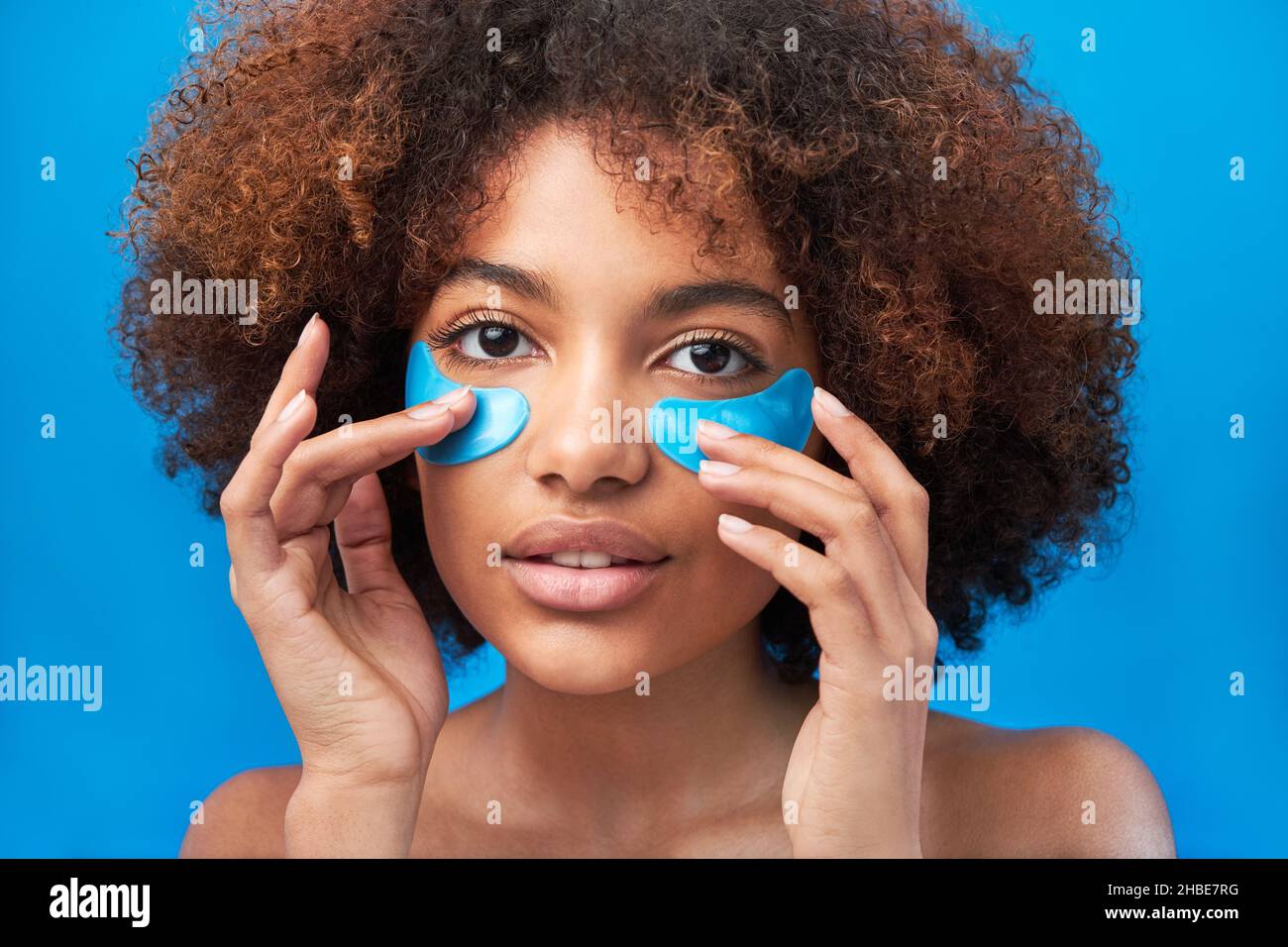Junge Frau mit langen lockigen Brünetten Haaren berührt blaue Schönheitsflecken gegen Beutel unter den Augen im Studio helle Wand aus der Nähe Stockfoto