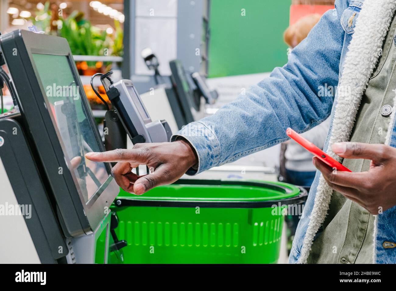 Der Finger eines Afrikaners in Nahaufnahme an der Supermarktkasse wählt das gewünschte Produkt auf dem elektronischen Bildschirm der Kasse mit einem Telefon in den Händen vor dem Hintergrund von grünen Einkaufskörben, Einzelhandel und Self-Service-Kasse in einem Hypermarkt aus Stockfoto