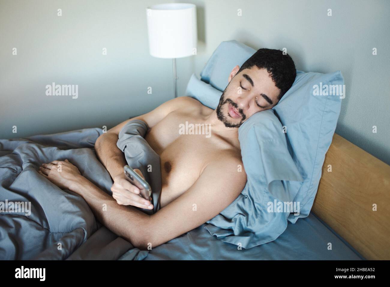 Ein Mann aus mehreren Rassen, ein bärtiger Mann, stand morgens auf den Kissen und überprüfte das Telefon im Bett, während er aufwacht. Bett mit blauen Laken und blauer Decke Stockfoto