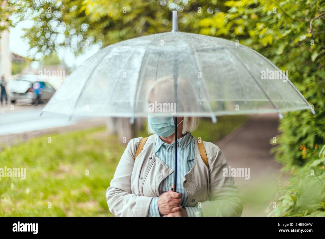 Porträt einer erwachsenen Frau in einem weißen Hut und einem warmen Schal unter einem transparenten Regenschirm in einem Herbstpark im Freien, an kalten Regentagen Stockfoto