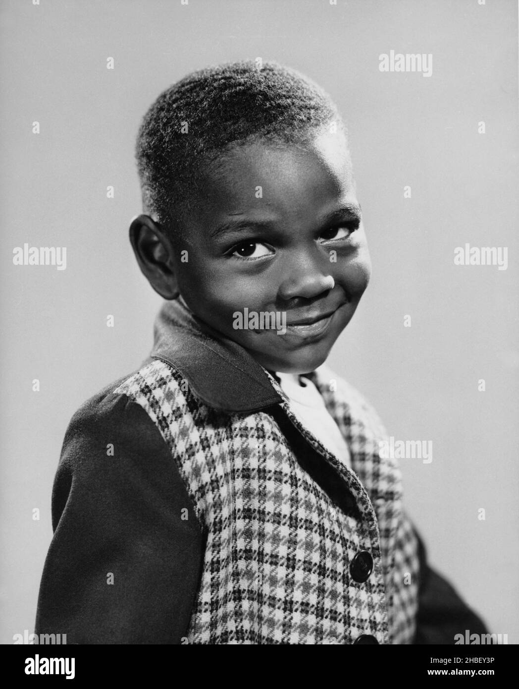 Aftrikanisch-amerikanischer Junge, der seitwärts sitzt und mit einem halblächelnden Lächeln auf die Kamera zurückblickt. Ca. 1965 Stockfoto