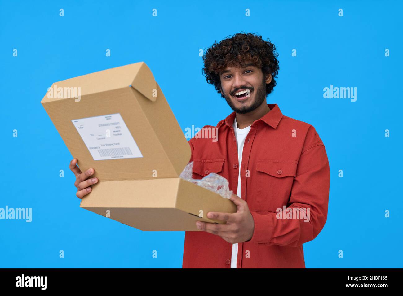Glücklicher indischer junger Mann mit offenem Paketkasten isoliert auf blauem Hintergrund. Stockfoto