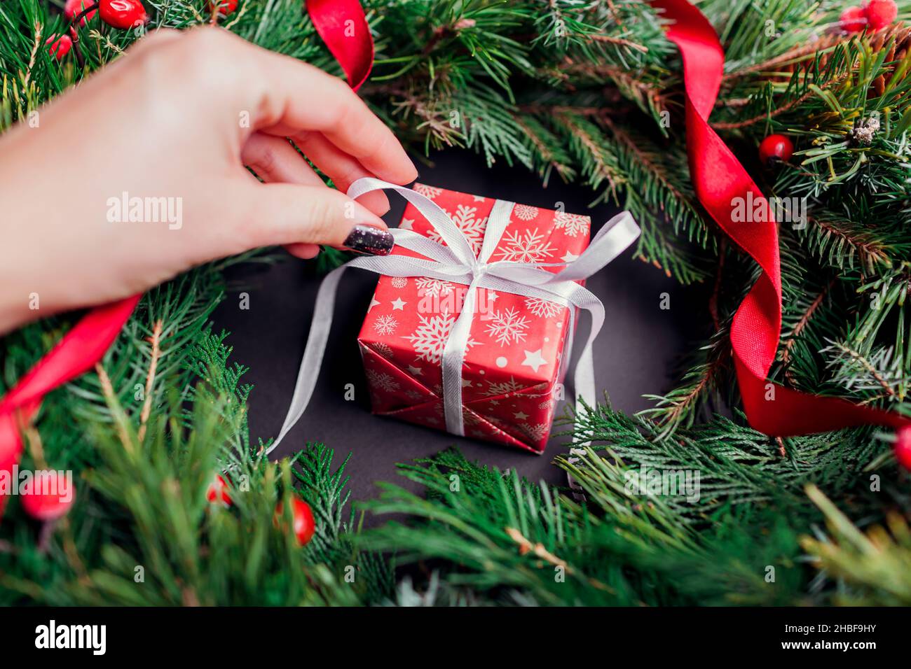 Frau öffnet Weihnachts- oder Neujahrsgeschenk-Box in festliches Papier und Band in Tannenkranz verpackt. Präsent für den Winterurlaub Stockfoto