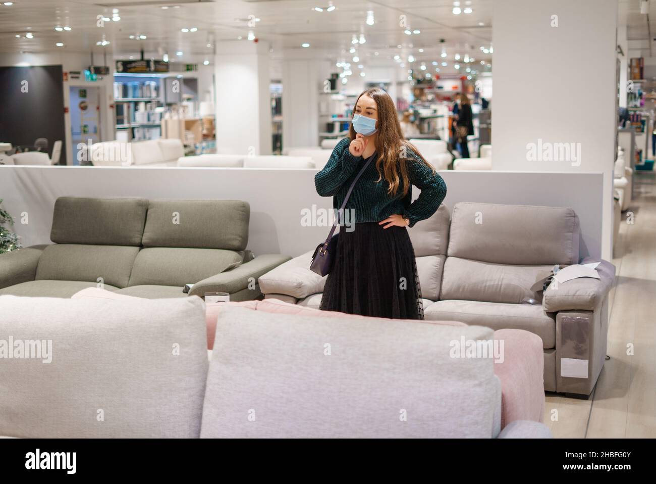 Junge nachdenkliche Frau in Gesichtsmaske, die vor der Couch im Möbelgeschäft steht und ihr die Hand auf das Kinn legt, untersucht nachdenklich das neue Sofa, während sie darüber nachdenkt, es für das Wohnzimmer zu Hause zu kaufen Stockfoto