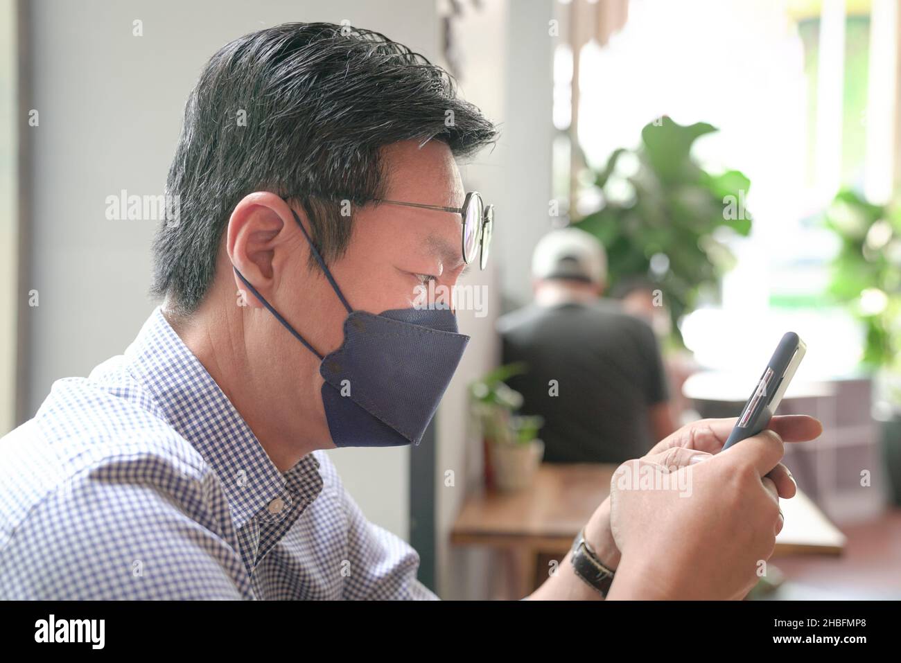 Asiatischer Mann mit Gesichtsmaske, der sein Telefon benutzt, SMS in einem Restaurant. Stockfoto