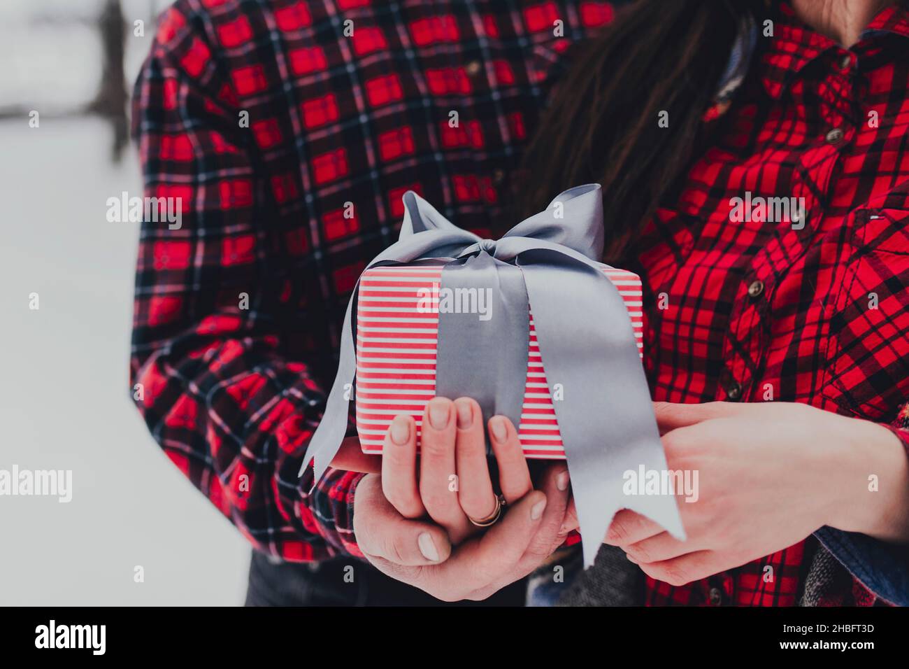 Freund gibt eine Geschenkbox Geschenk mit Band im Freien aufgrund der heiligen Valentinstag zu ihrer Freundin. Verschneite Wälder, Bäume mit Schnee. Zwei sind in roter Kleidung gekleidet. Schönes Paar liebt sich gegenseitig Stockfoto