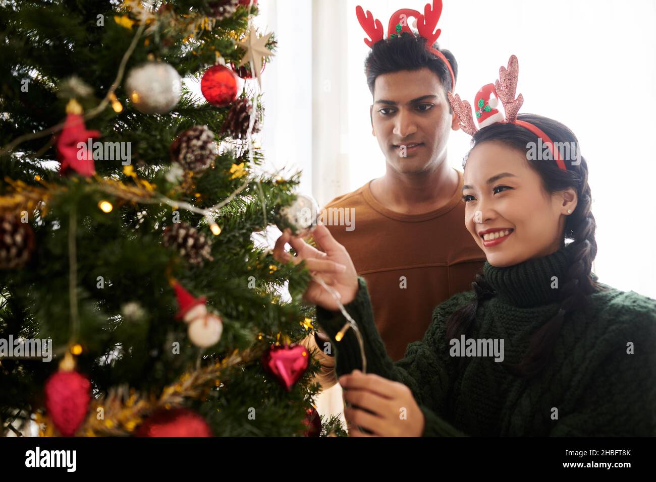 Glückliches devirse junges Paar, das Weihnachtsbaum zu Hause mit bunten Kugeln schmückt Stockfoto