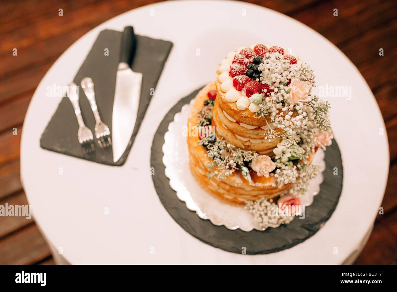 Hochzeitstorte mit Himbeeren, Heidelbeeren und Blumen auf einem Teller auf einem Tisch mit Tischdecke Stockfoto