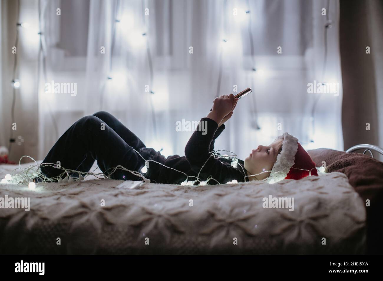 Kleinkind mit niedlichem weihnachtsmütze, im Bett mit Lichtern liegend, Weihnachtsmusik auf dem Mobiltelefon hörend Stockfoto