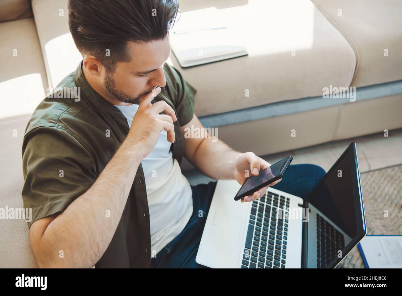 Kaukasischer Mann mit Mobiltelefon, während er zu Hause mit einem Laptop auf dem Boden sitzt. Geschäftsmann mit Laptop. Unternehmenstechnologie. Internet Stockfoto