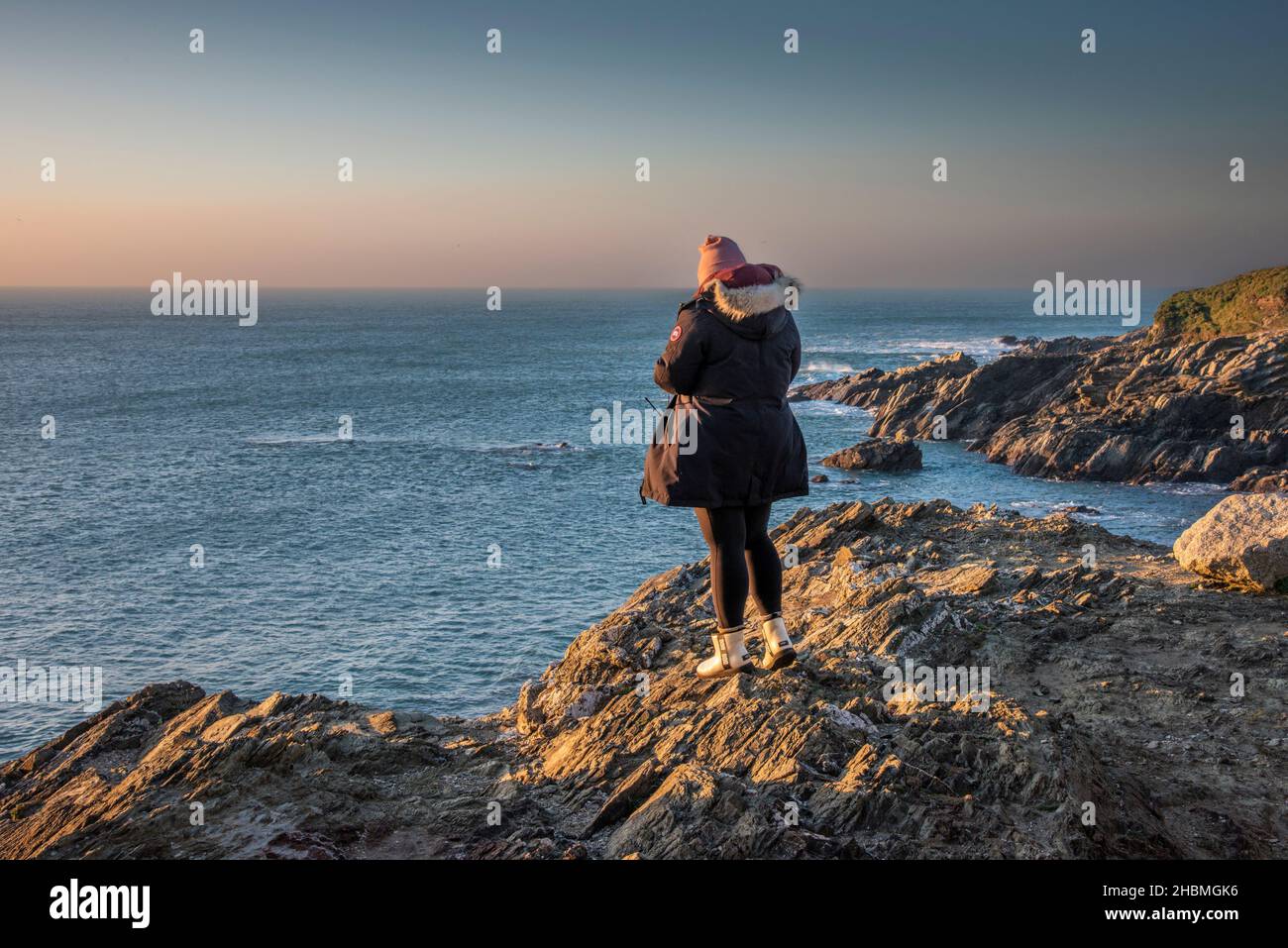 Eine Frau in warmer Kleidung gegen das kalte Wetter, die einen spektakulären Sonnenuntergang an der Küste von North Cornwall erlebt. Stockfoto