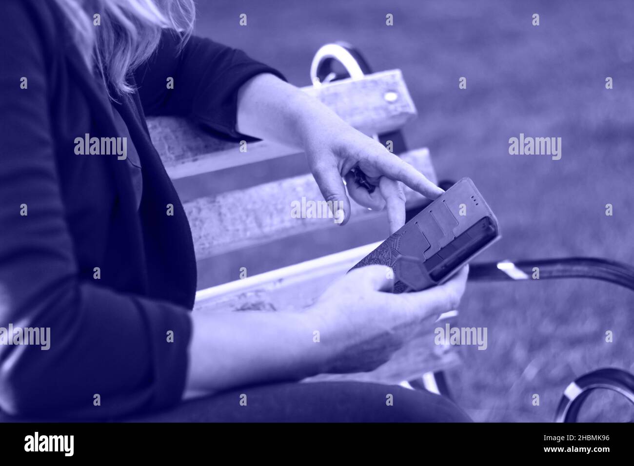 Unschärfe-Unschärfe-Nahaufnahme weibliche Hände suchen auf dem Telefon außerhalb, im Freien. Frau in lila Bluse mit Smartphone, während sie auf einer Bank im Park sitzt. Farbe Stockfoto