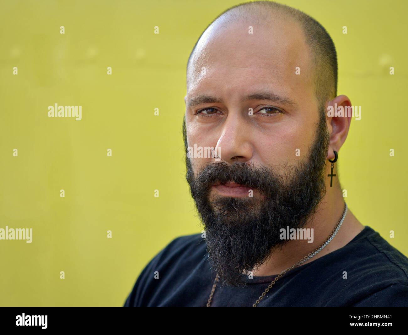 Ein gutaussehender maskuliner, einfallsreicher junger kaukasischer Mann mit hoher Stirn, schwarzem Bart und christlichem Kreuzohrring blickt den Betrachter streng an. Stockfoto