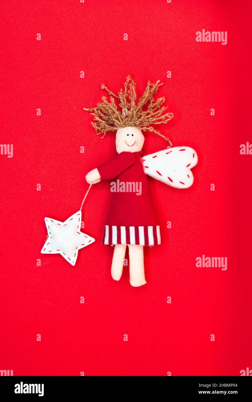 Niedlicher Weihnachtsengel aus Stoff isoliert. Komposition aus lächelnder Puppe mit Flügeln in rotem Kleid und Stern auf rotem Hintergrund. Stockfoto