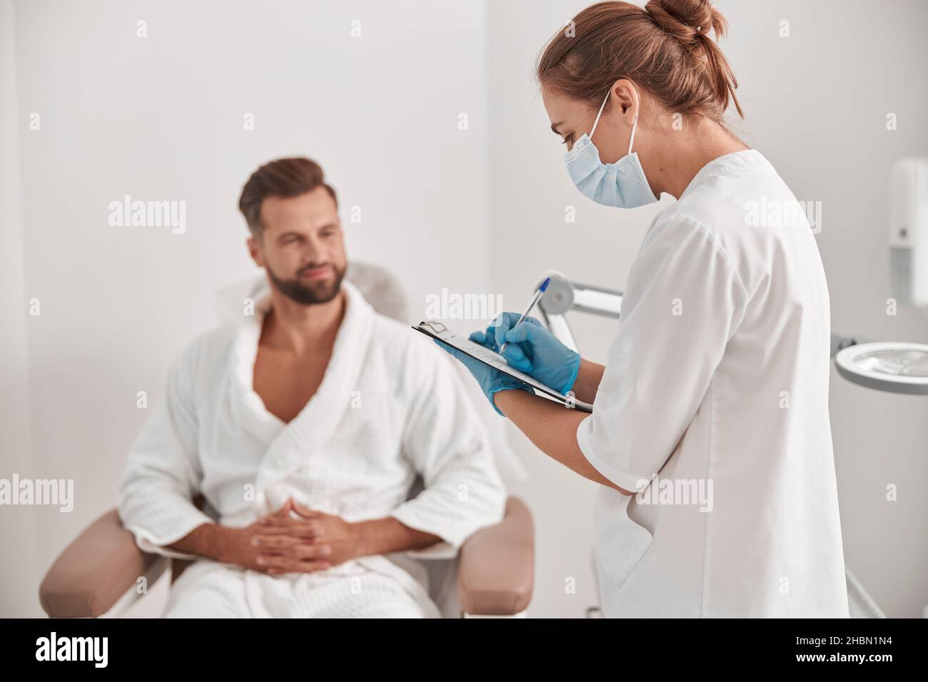Administrator in Maske und Handschuhe füllt Form Untersuchung männlichen Patienten in der Kosmetologie Klinik Stockfoto