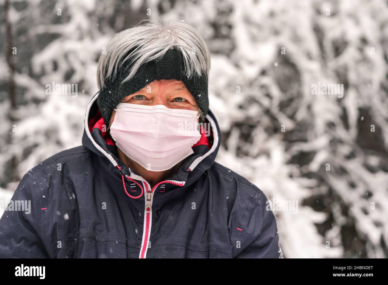 Ältere ältere Frau mit grauen Haaren, die eine rosa Einweg-Virus-Gesichtsmaske trägt, verschwommener Winter-Schnee bedeckte Bäume im Hintergrund Stockfoto