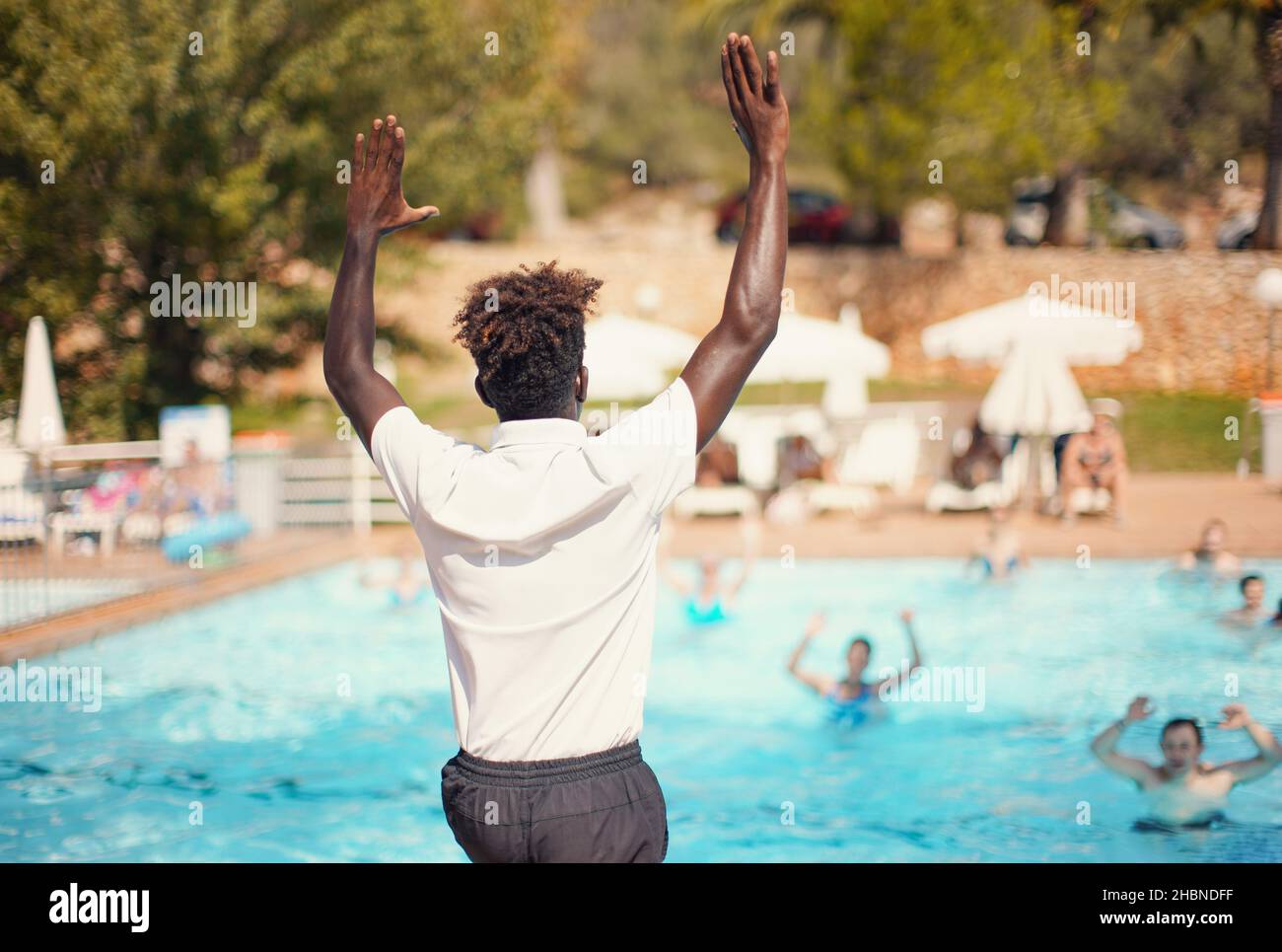 Der schwarze, sportliche Animator des Hotelresort trainiert vor dem Pool, Blick von der Seite hinter dem Hintergrund der verschwommenen Menschen - Wasseraerobic-Aktivitäten Stockfoto