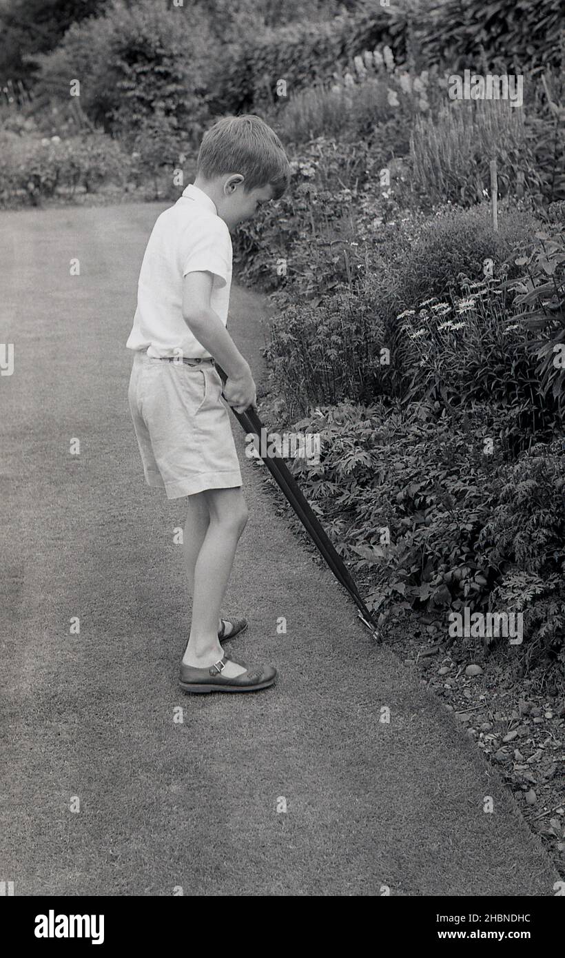 1950s, historisch, draußen in einem Garten, ein Junge in kurzen Hosen, Kurzarmhemd und Sandalen, der bei der Gartenarbeit hilft, den Rand eines Rasens mit einem Paar lang gehandelte Scheren oder Scheren trimmt, England, Großbritannien. Stockfoto