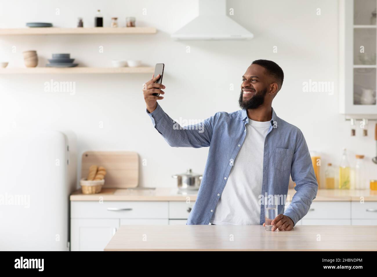 Glücklicher junger schwarzer Kerl mit Glas sauberem Wasser macht Selfie, Videoanruf in skandinavischer Kücheneinrichtung Stockfoto