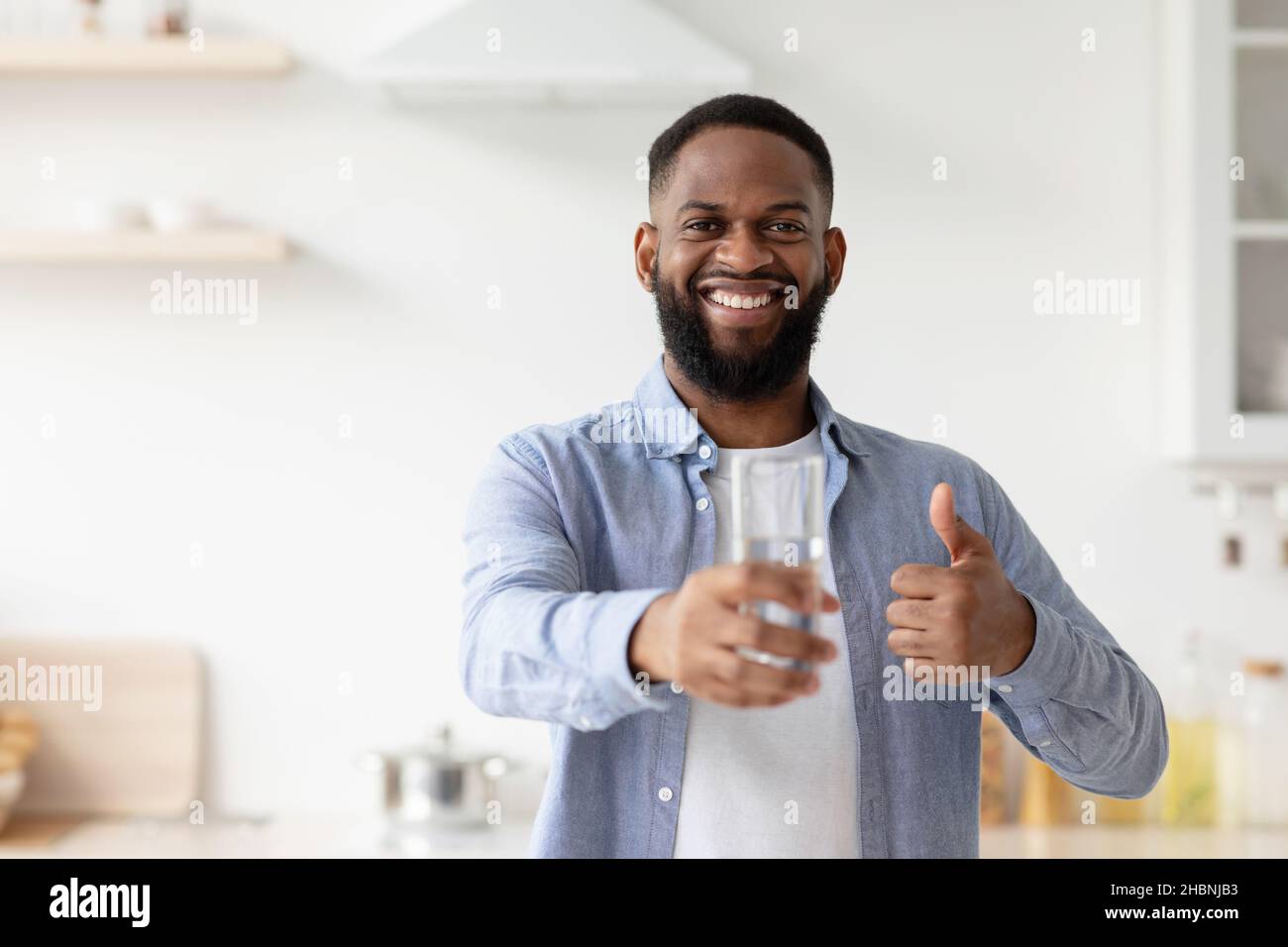 Lächelnder, tausendjähriger schwarzer Kerl zeigt ein Glas Wasser in der Hand, Daumen hoch im Kücheninnenraum Stockfoto