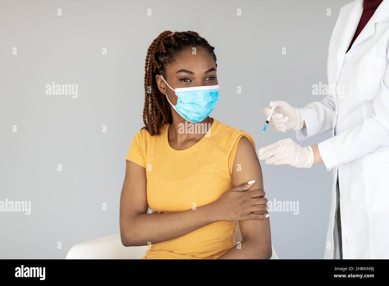 Ärztin, die der jungen schwarzen Frau in medizinischer Maske, grauem Studiohintergrund, eine Injektion von Anti-Covid-Impfstoff macht Stockfoto