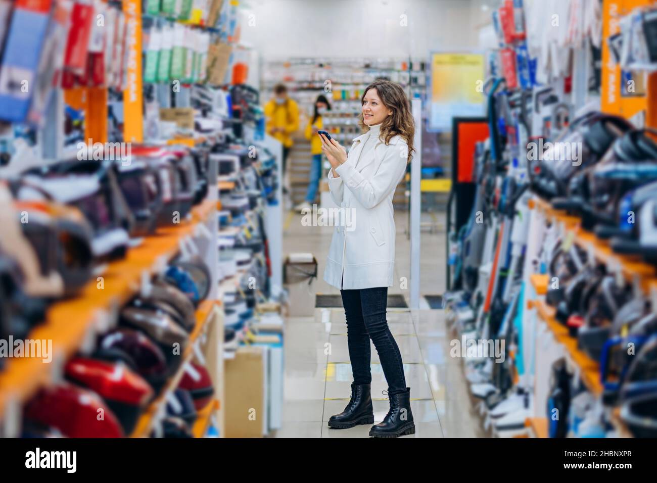 Lächelnde, moderne Frau in einem weißen Mantel wählt Haushaltsgeräte mit einem Telefon in den Händen in einem Hypermarkt Stockfoto