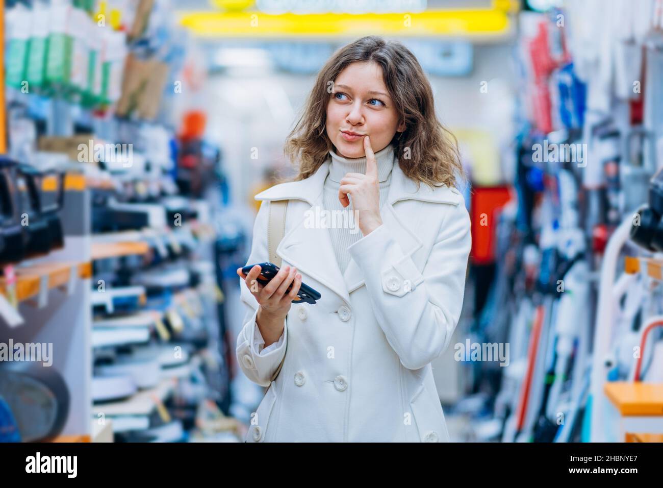 Die schöne Kauffrau in einem weißen Mantel dachte mit einem Telefon in der Hand in einem Haushaltswarenladen in einem Supermarkt, an die Emotion der Freude und die Schwierigkeit, sich vor dem Kauf zu entscheiden Stockfoto