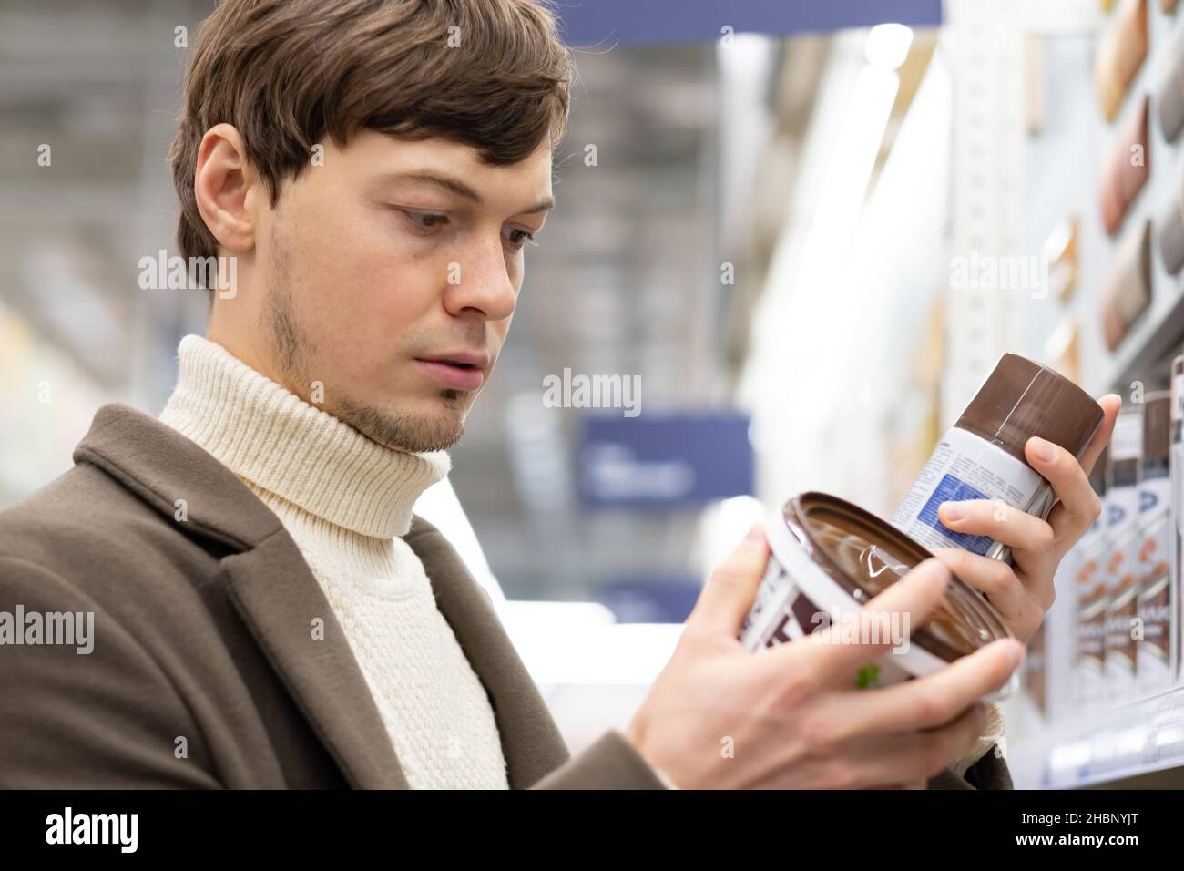 Porträt eines europäischen Mannes in einem Mantel wählt Farbe in einem Baumarkt zur Reparatur, liest sorgfältig die Komposition Stockfoto