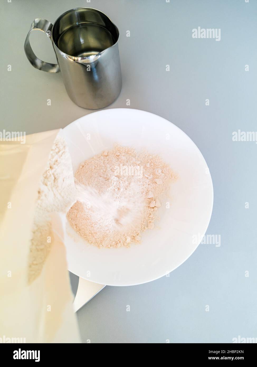 Mehl wird aus einem Beutel in eine Schüssel neben einem Metallbecher mit Wasser gegießt Stockfoto