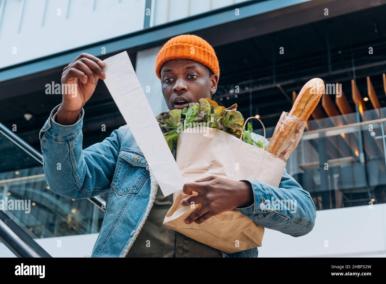 Zweifelhaft afroamerikanische Person in Denim Jacke schaut auf Verkaufspapier Quittung Gesamthaltepaket mit Lebensmitteln auf Rolltreppe Stockfoto