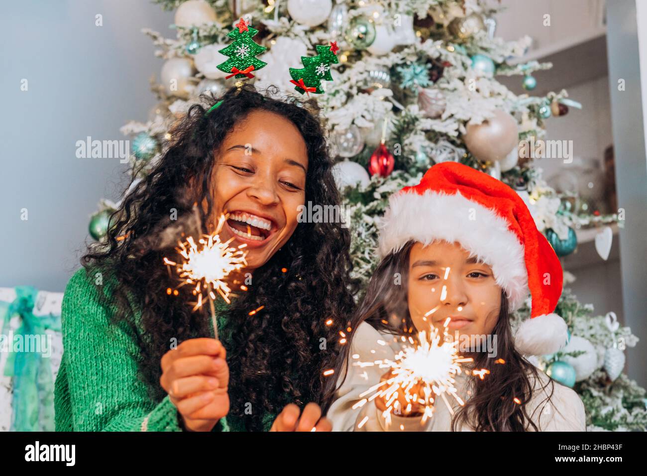 afroamerikanische Mutter und kleine Tochter in einem weihnachtshut mit Wunderkerzen und Lachen vor dem Hintergrund eines weihnachtsbaums Stockfoto