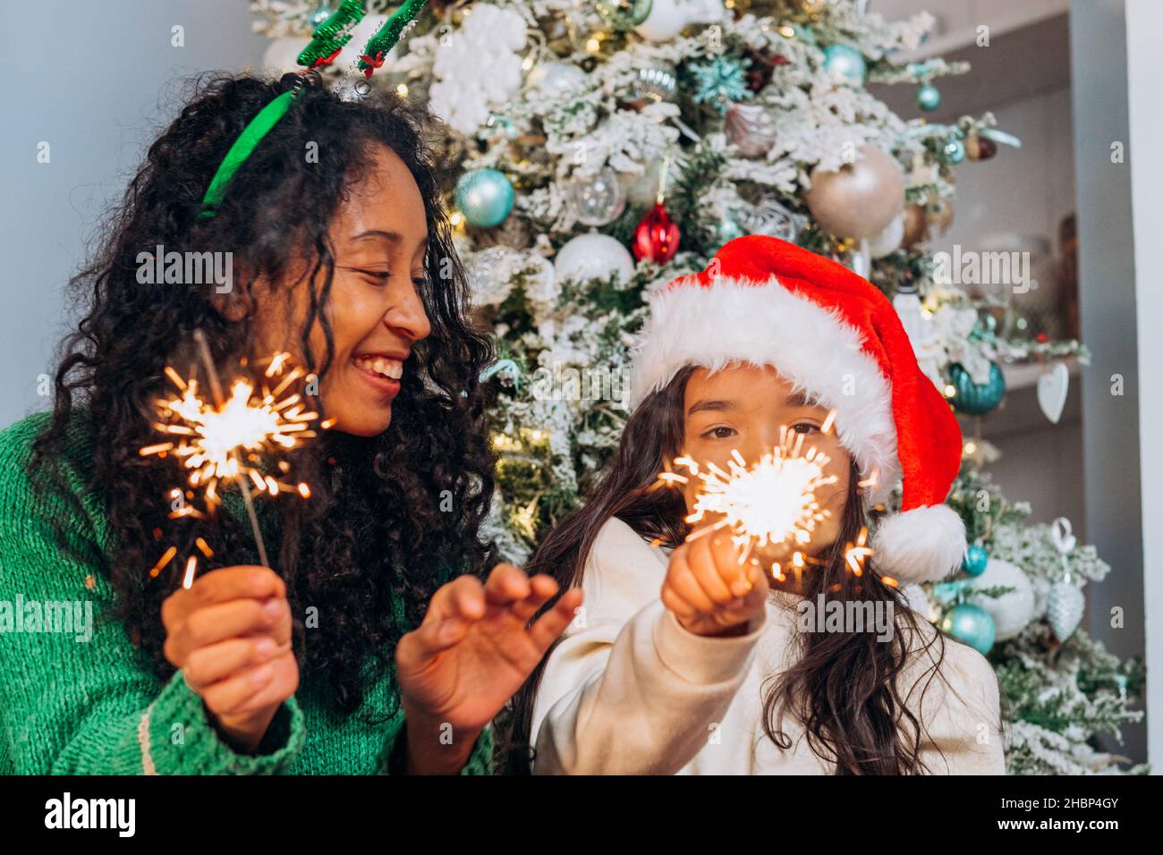 afroamerikanische Mutter und kleine Tochter in einem weihnachtshut mit Wunderkerzen und Lachen vor dem Hintergrund eines weihnachtsbaums Stockfoto