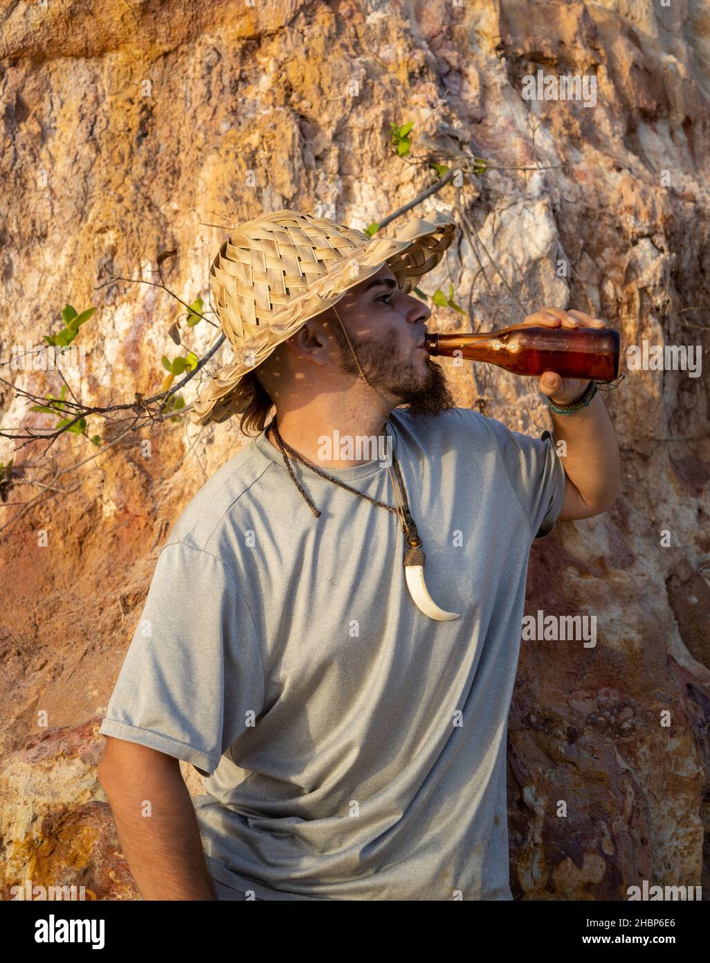 Junger Mann in einem rustikalen Hut, der ein Bier am Strand trinkt. Stockfoto