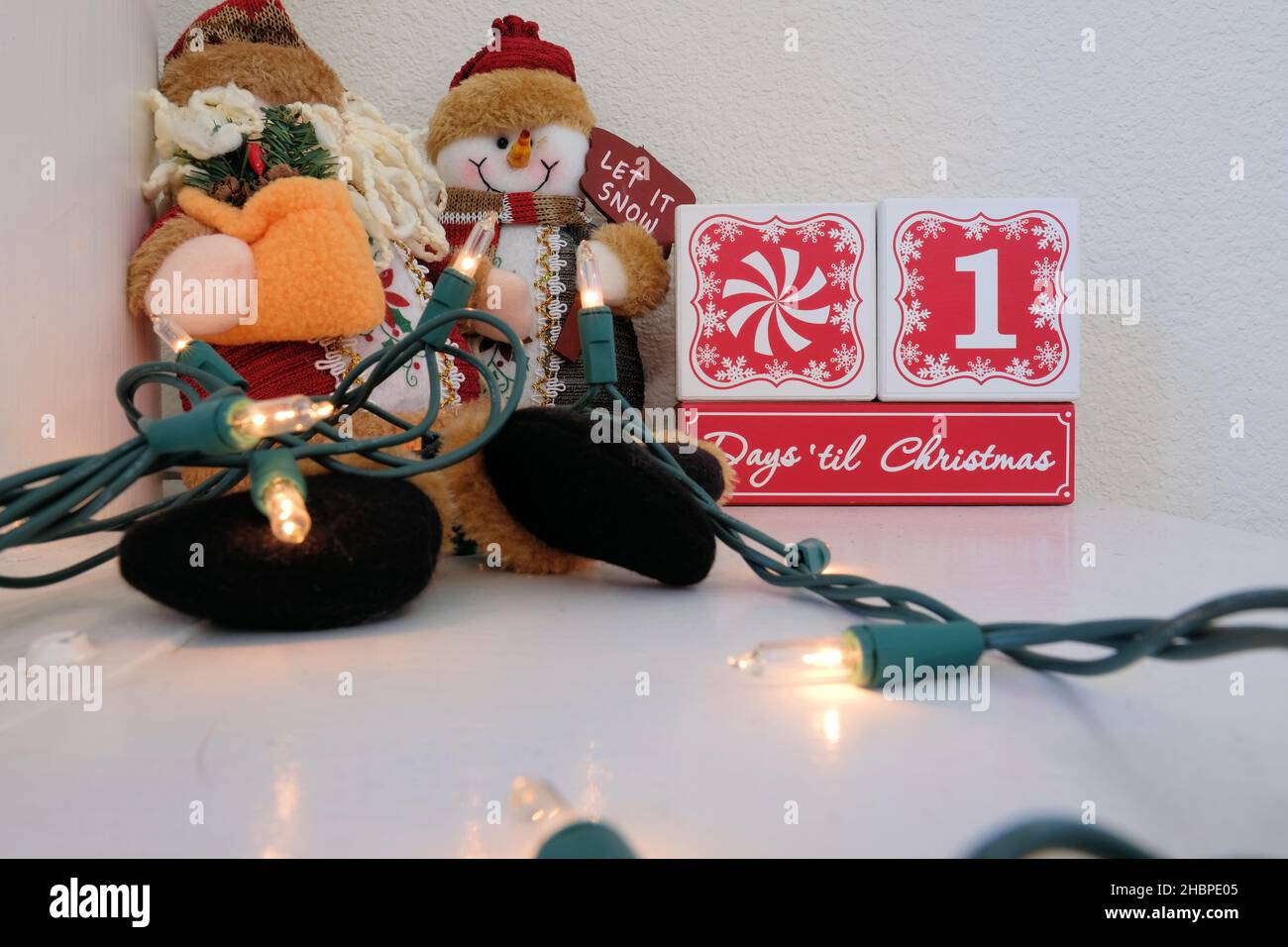 Eines Tages bis Weihnachten Countdown-Kalender aus Holzblöcken auf einem Mantel mit Lichtern und einem Plüsch Spielzeug Schneemann. Stockfoto