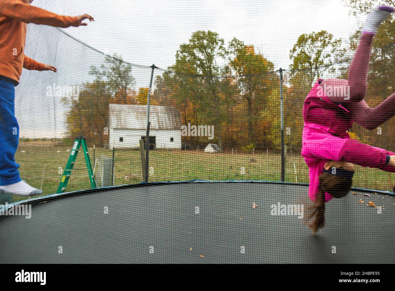 Ein akrobatisches junges Mädchen dreht sich auf den Kopf, als zwei Kinder auf einem Trampolin auf ihrer Familienfarm in der Nähe von Spencerville, Indiana, USA, springen. Stockfoto