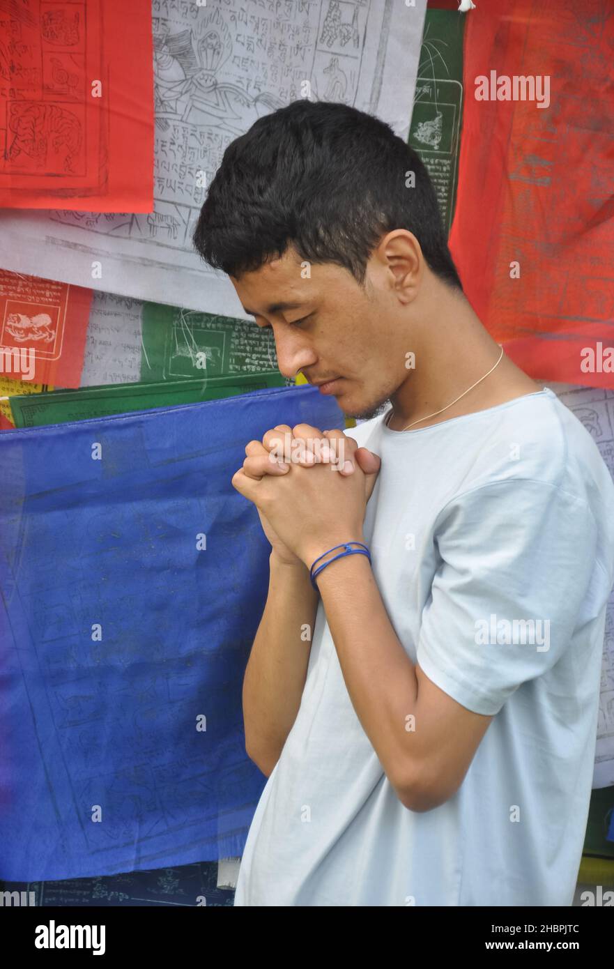 Seitenansicht eines treuen jungen Mannes, der mit gefalteten Händen betet, Gott mit dem Kopf nach unten anbetet und gegen buddhistische Gebetsfahnen steht Stockfoto