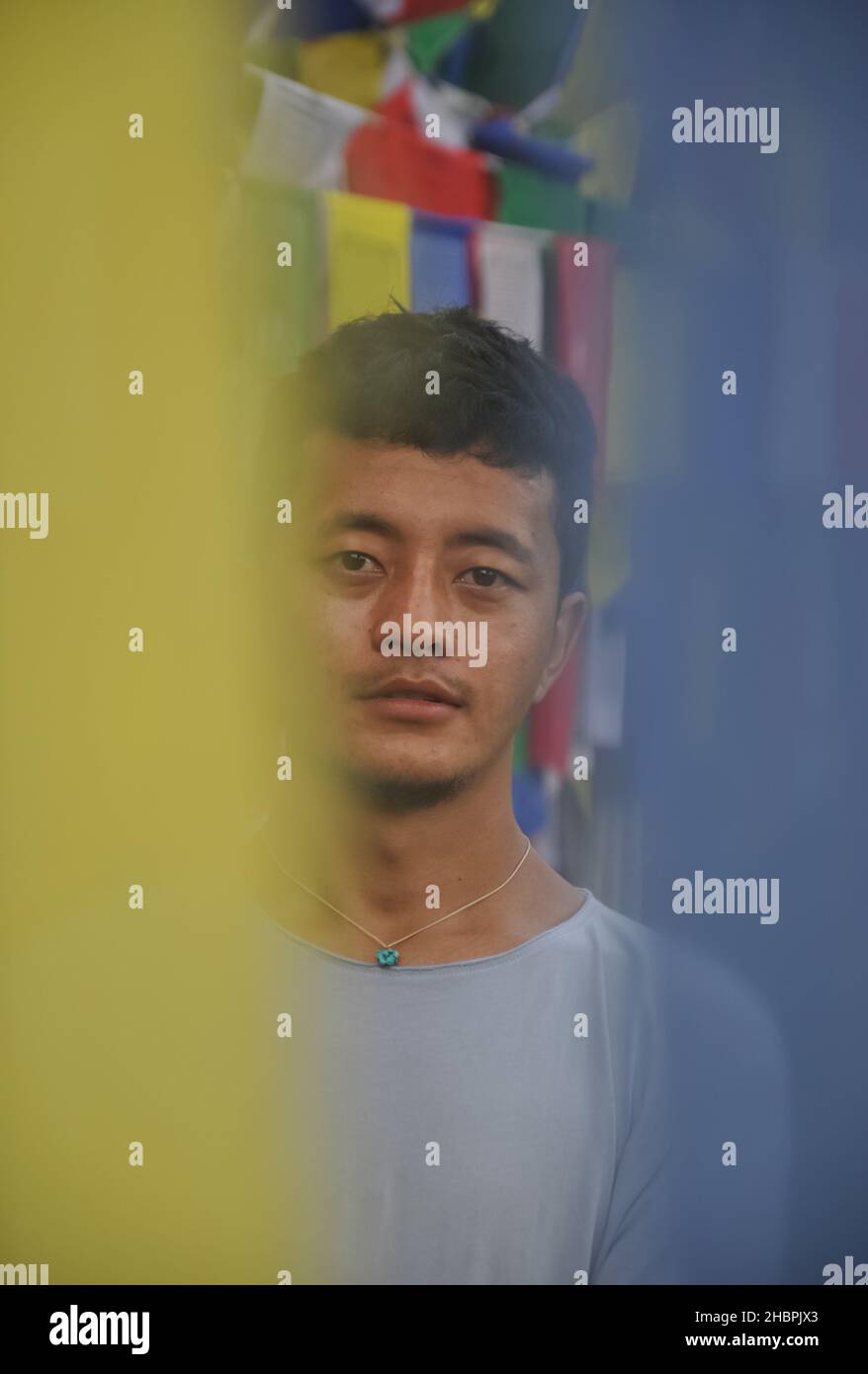 Kreatives Foto eines asiatischen jungen Mannes, der die Kamera anschaut, während er gegen buddhistische Gebetsfahnen steht Stockfoto