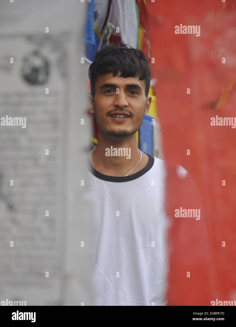 Selektiver Fokus eines jungen indischen Mannes, der die Kamera anschaut, während er um buddhistische Gebetsfahnen steht Stockfoto