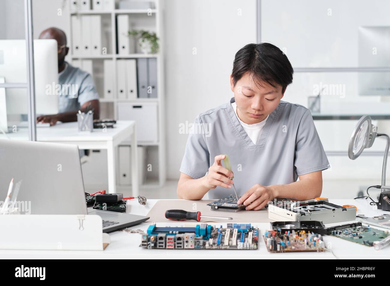 Junge Chinesin in Uniform, die kleine Details der Leiterplatte fixiert, während sie am Schreibtisch gegen ihren afrikanischen männlichen Kollegen sitzt Stockfoto