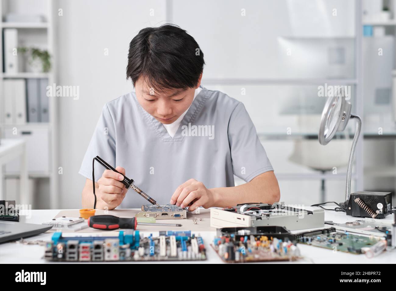 Junge chinesische Technikerin in Uniform Reparatur Platine mit Mikroprozessor während sitzen am Arbeitsplatz Stockfoto