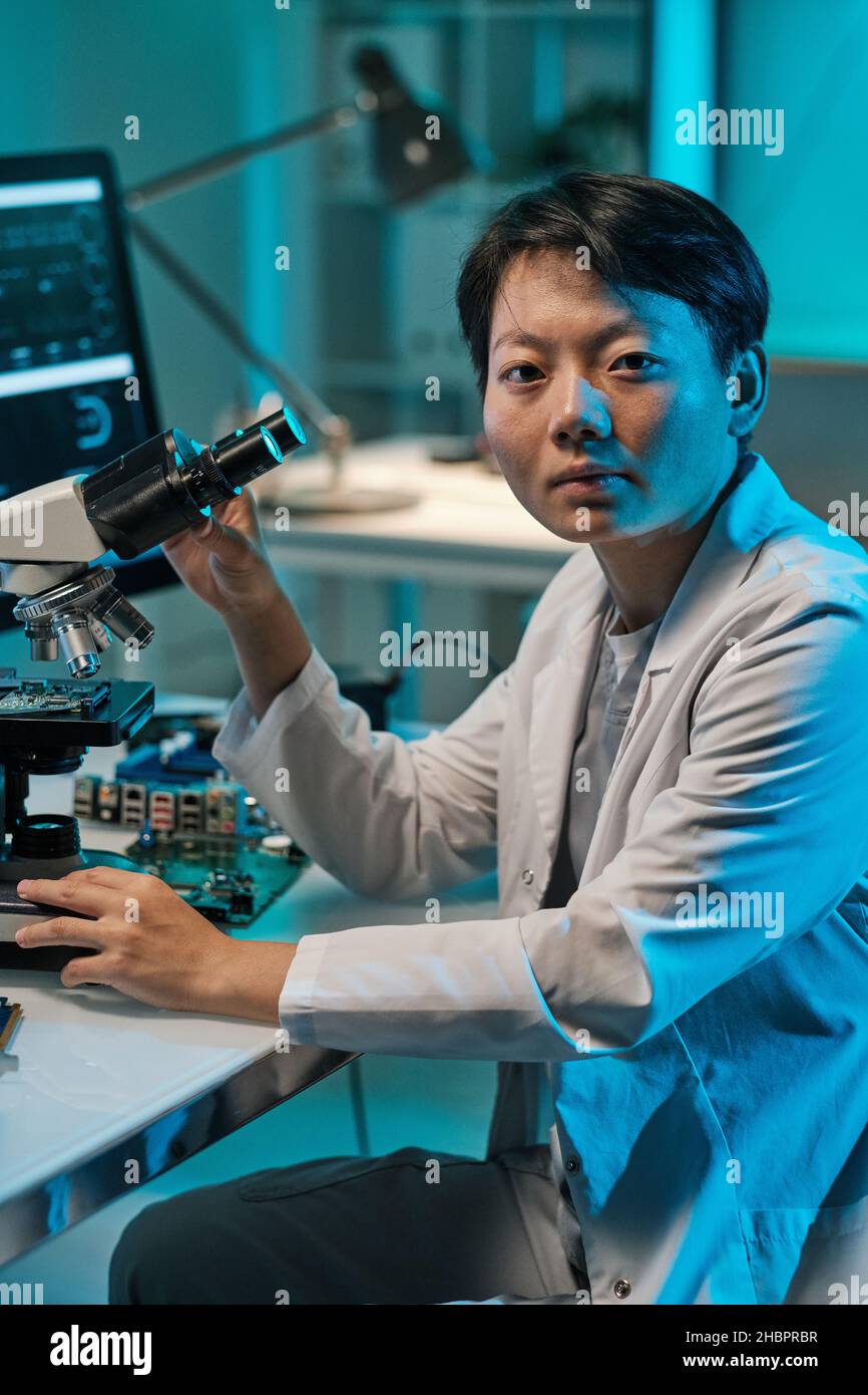 Junge Chinesin im Weißkochfeld, die während der Arbeit mit dem Mikroskop im technischen Labor auf die Kamera schaut Stockfoto