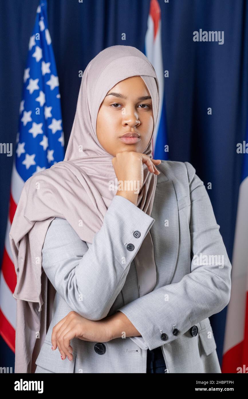 Junge ernsthafte Politikerin in Hijab und formalwear, die Sie anschaut, während sie gegen Flaggen mehrerer Länder steht Stockfoto