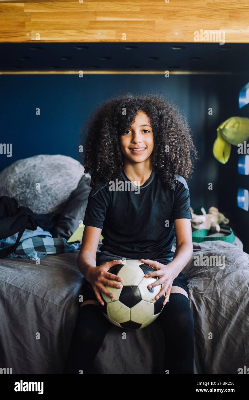 Portrait von lockiges Haar Mädchen mit Fußball sitzt auf dem Bett zu Hause Stockfoto