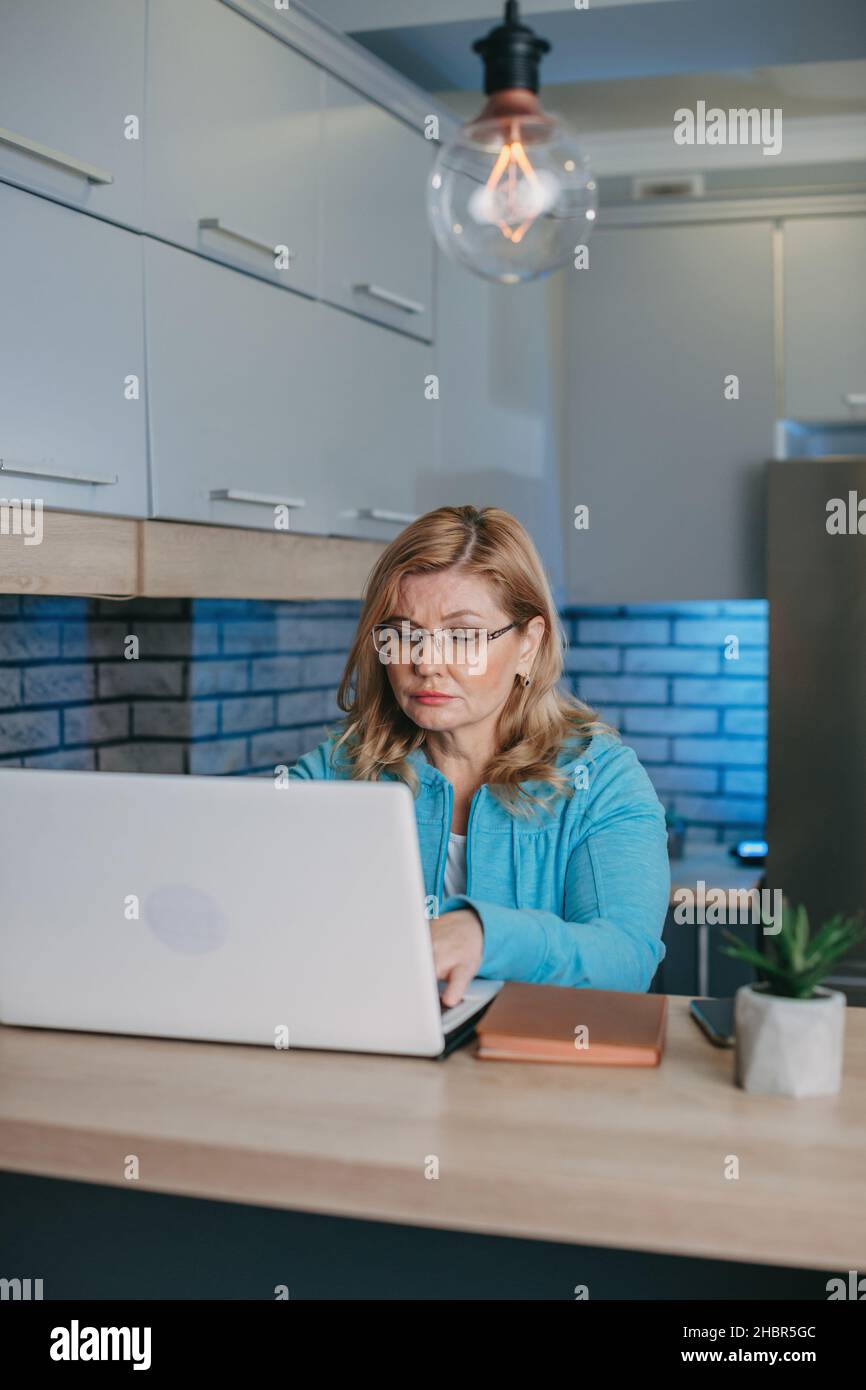 Aufnahme einer fokussierten Geschäftsfrau mittleren Alters in einer Brille, die vor dem Laptop sitzt und arbeitet. Quarantäne-Lebensstil. Blonde Frau. Stockfoto