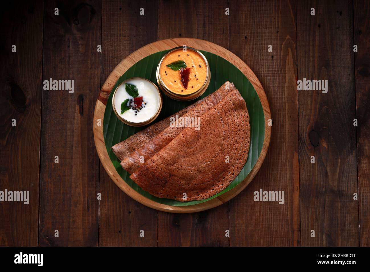 Ragi Dosa, ein gesundes südindisches Frühstücksartikel, das auf einem runden Holzsockel mit Bananenblatt und Kokosnuss-Chutney daneben angeordnet ist. Stockfoto