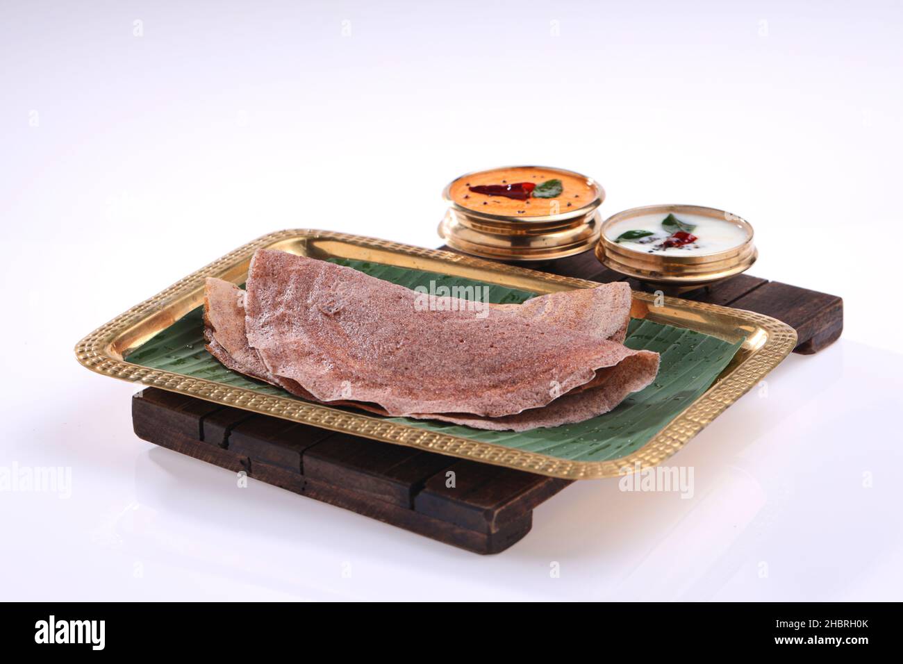 Ragi Dosa, ein gesundes südindisches Frühstückselement, das auf einem rechteckigen Messingteller mit Bananenblatt und Kokos-Chutneys daneben angeordnet ist. Stockfoto