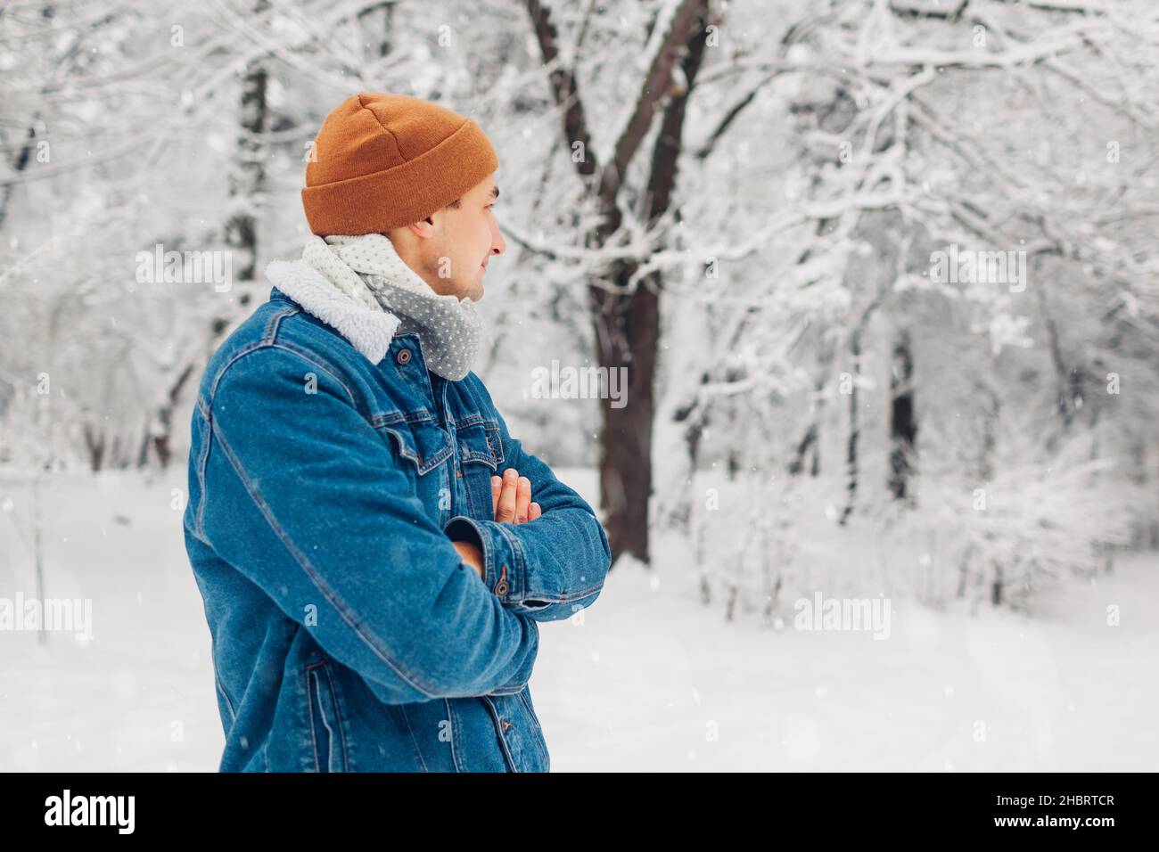 Portrait eines stilvollen Mannes im verschneiten Winterpark, der einen Jeansmantel mit gekreuzten Händen im Freien trägt. Hübscher Kerl genießt fallenden Schnee und Landschaft. Männlich Stockfoto